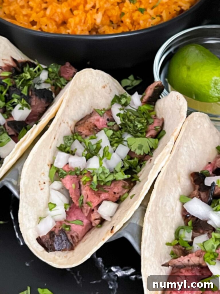 An overhead view of multiple flank steak street tacos with rice, chips, and lime wedges on a dark background, showcasing a complete meal.