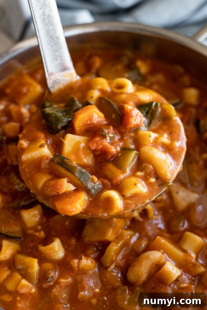 A soup ladle lifting a generous scoop of hearty minestrone soup, showing the rich broth, pasta, beans, and various vegetables, ready to be served into a bowl.