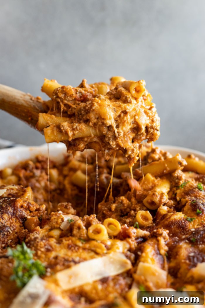 Pulling up a large spoonful of baked ziti from a white casserole dish, showcasing melted cheese and rich meat sauce.