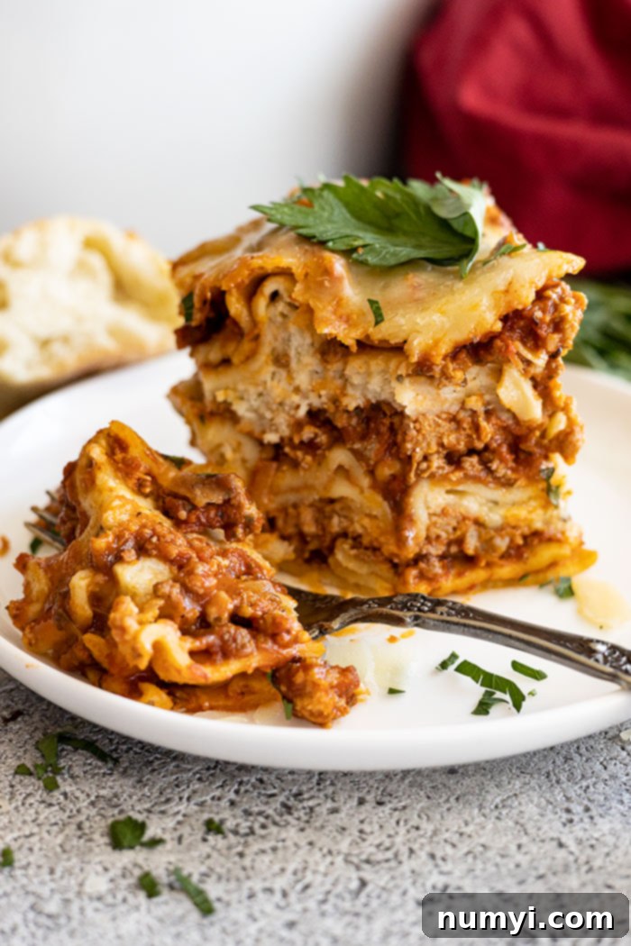 A tall slice of lasagna on a white plate with a bite taken out. Garnished with fresh parsley and parmesan cheese.