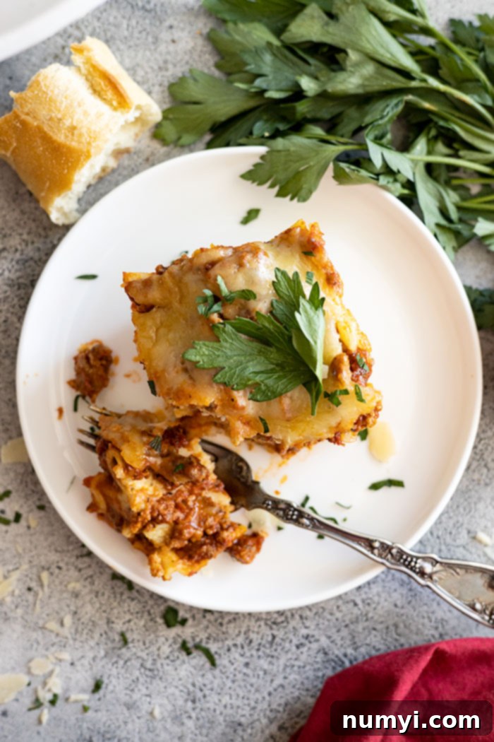 Overhead view of a piece of lasagna on a white plate with bread and parsley off to the side and a red napkin in the corner.