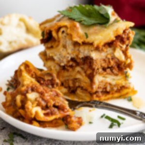 A tall slice of lasagna on a white plate with a bite taken out. Garnished with fresh parsley and parmesan cheese.