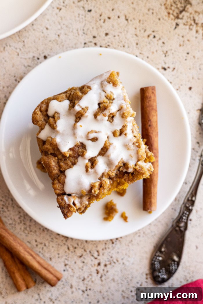 Overhead view of a delicious piece of pumpkin coffee cake with crumb topping on a white plate, with a fork and cinnamon sticks to the side.
