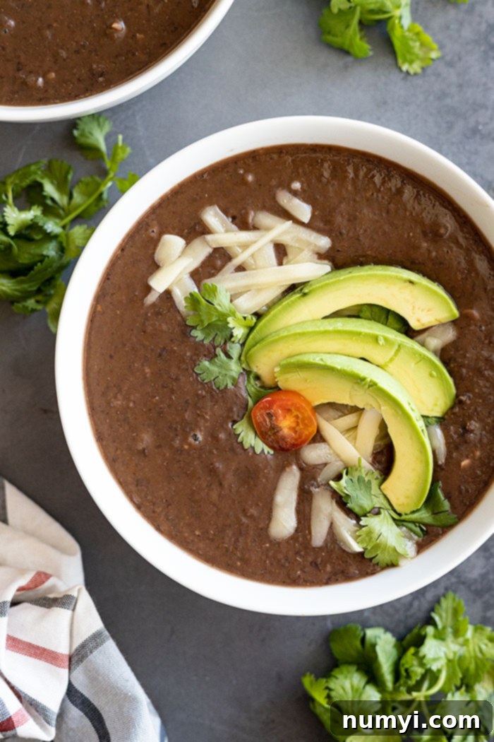 Hearty Black Bean Soup 2 Overhead view of black bean soup in a white bowl garnished with avocado, cheese, cilantro, and tomato.