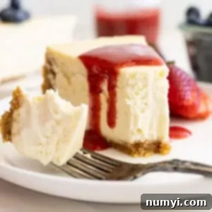 Straight on view of vanilla cheesecake on a white plate, strawberry sauce over the top, and a bite taken out showing how creamy and smooth it is.