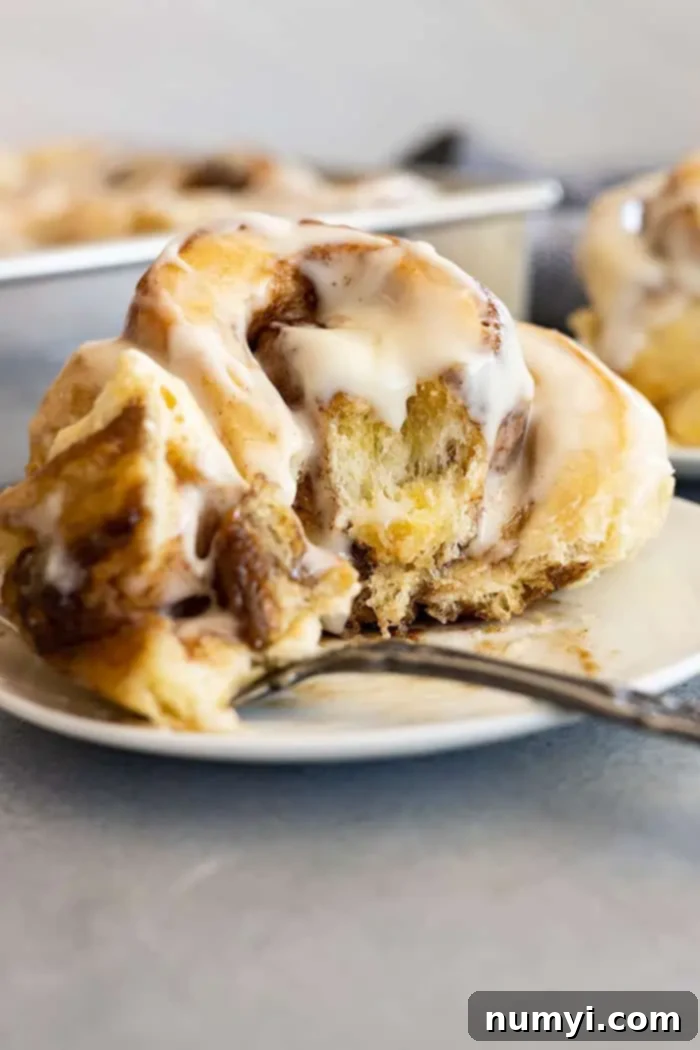 A perfectly baked, frosted cinnamon roll on a white plate with a bite taken, featuring a fork to the side, highlighting its soft texture and generous filling.