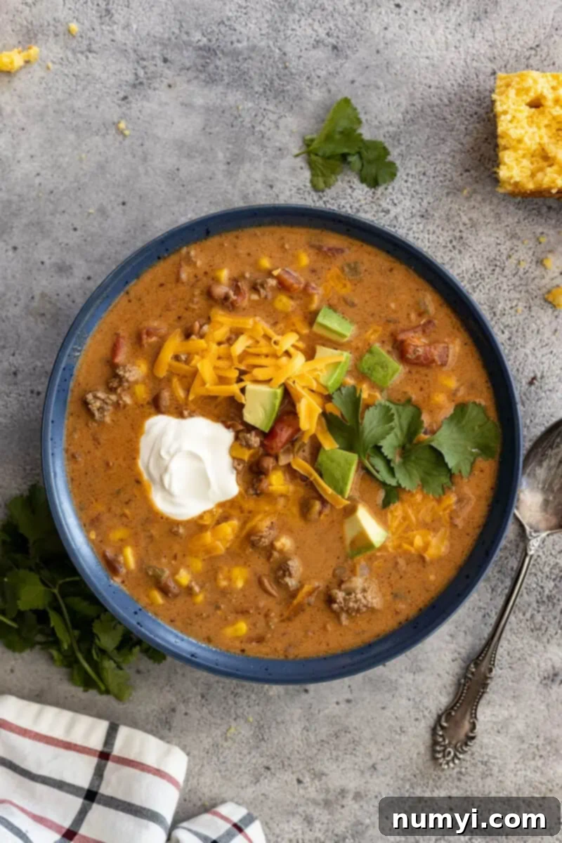Overhead view of a steaming bowl of creamy taco soup generously garnished with a swirl of sour cream, fresh avocado slices, bright green cilantro, and a sprinkle of grated cheddar cheese. It looks incredibly appetizing and ready to eat.