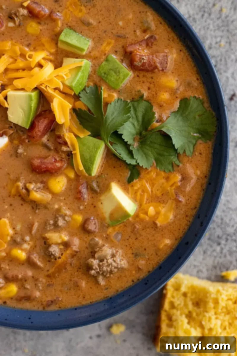 Close-up of a generous bowl of creamy taco soup, beautifully garnished with an assortment of fresh toppings including a dollop of sour cream, finely chopped cilantro, and a sprinkle of vibrant shredded cheese. A side of golden cornbread is visible in the background, suggesting a perfect pairing for this comforting meal.