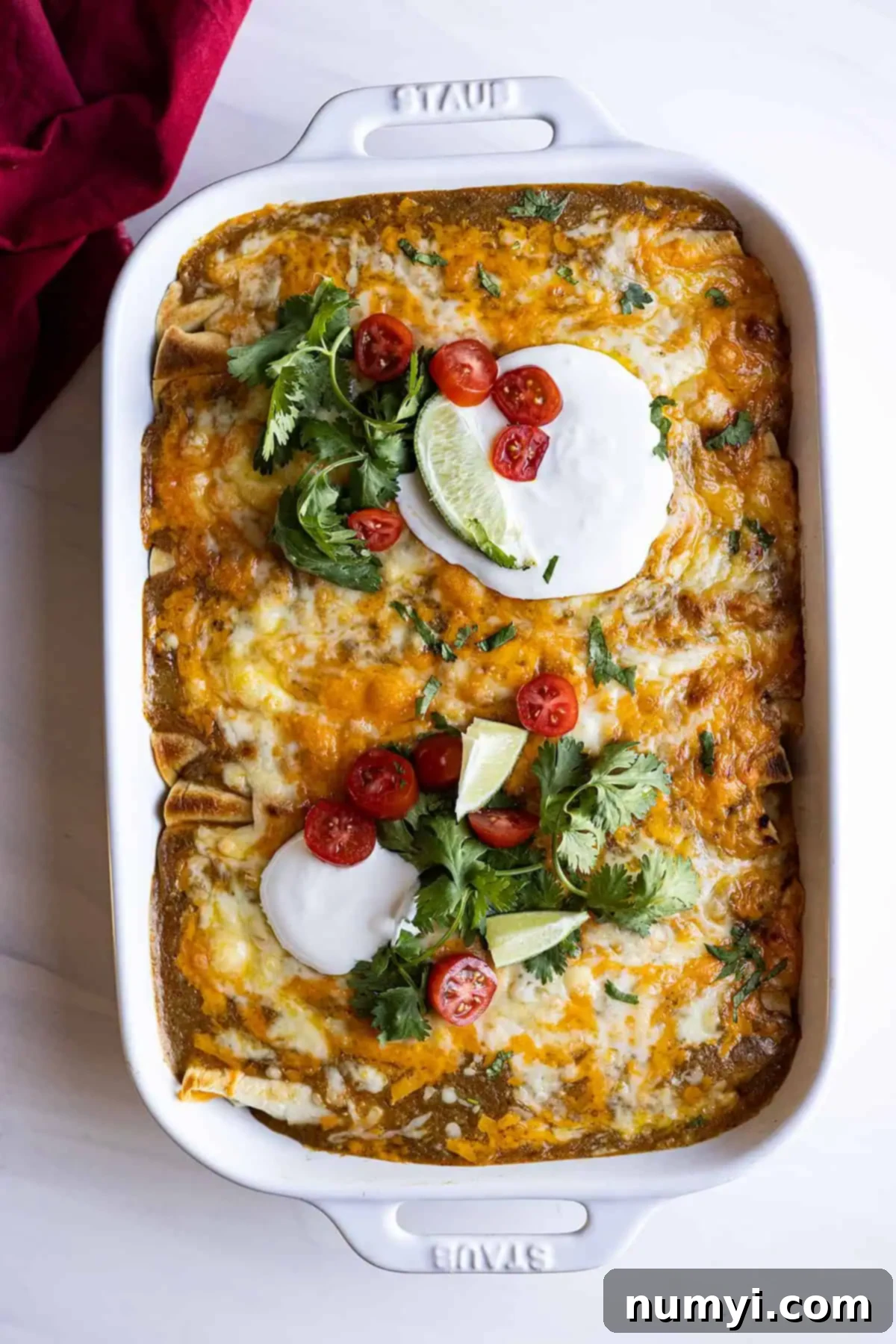 Overhead view of chicken enchiladas in a white casserole dish and garnished with sour cream, cilantro, tomatoes, and lime wedges. 