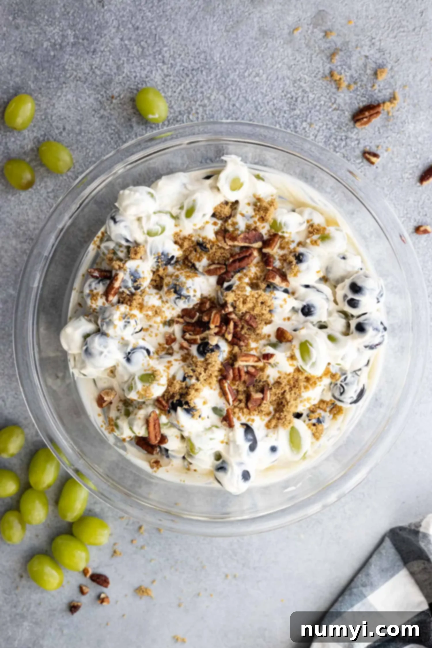 Overhead view of creamy grape salad in a bowl garnished with chopped nuts and brown sugar. Grapes and nuts sprinkled around the bowl.