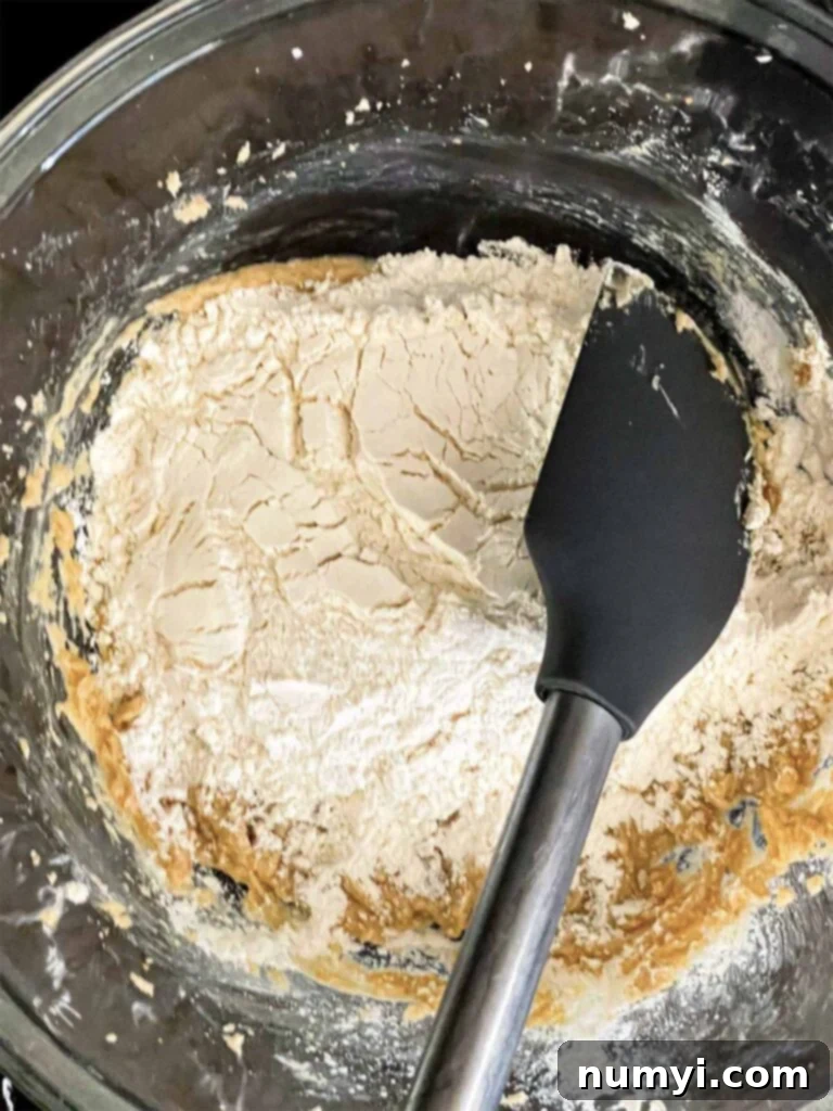 Dry ingredients being added to the wet ingredients in a glass mixing bowl for almond joy cookies, showing the flour being slowly incorporated.