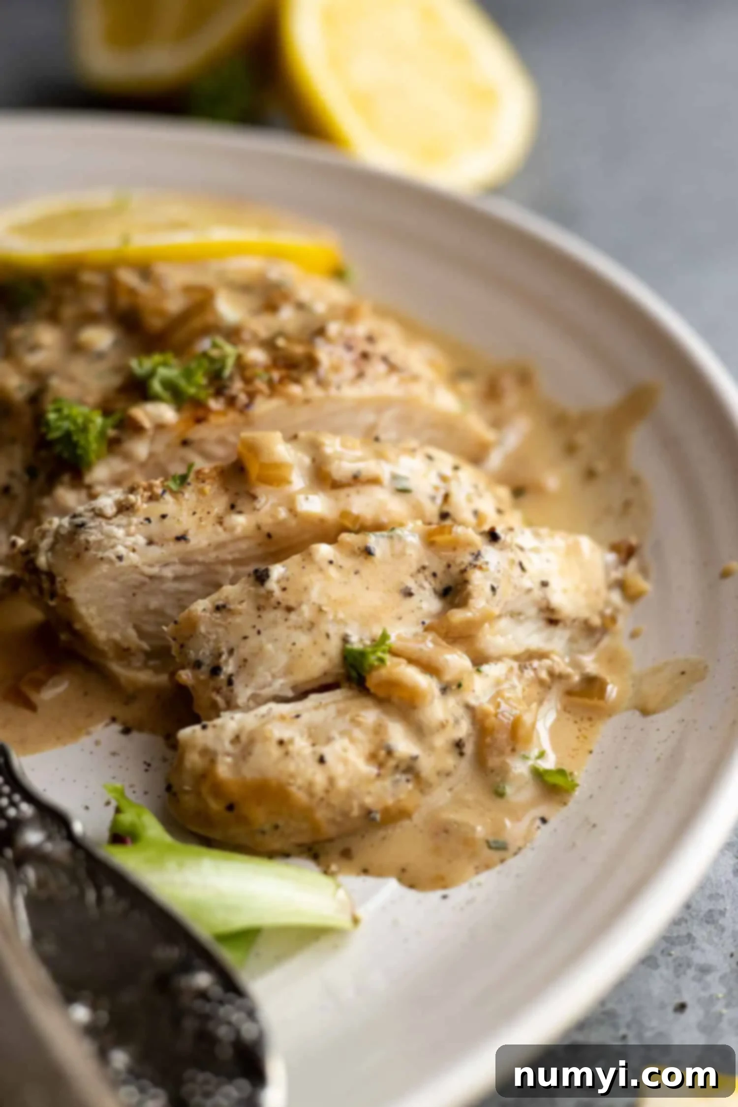 Close-up of creamy lemon chicken sliced on a plate, with a fresh cut lemon and sprig of rosemary in the soft background.