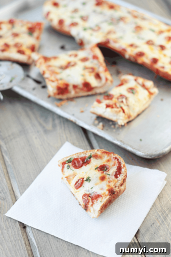 in the foreground is a pice of french bread pizza on a white napkin while the rest of the pizza is cut into pieces on a baking sheet in the background