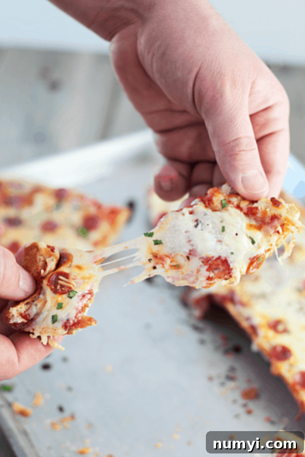 hands pulling a piece of this homemade french bread pizza recipe apart with the cheese stringing between both pieces and the remaining pizza on a baking sheet in the background