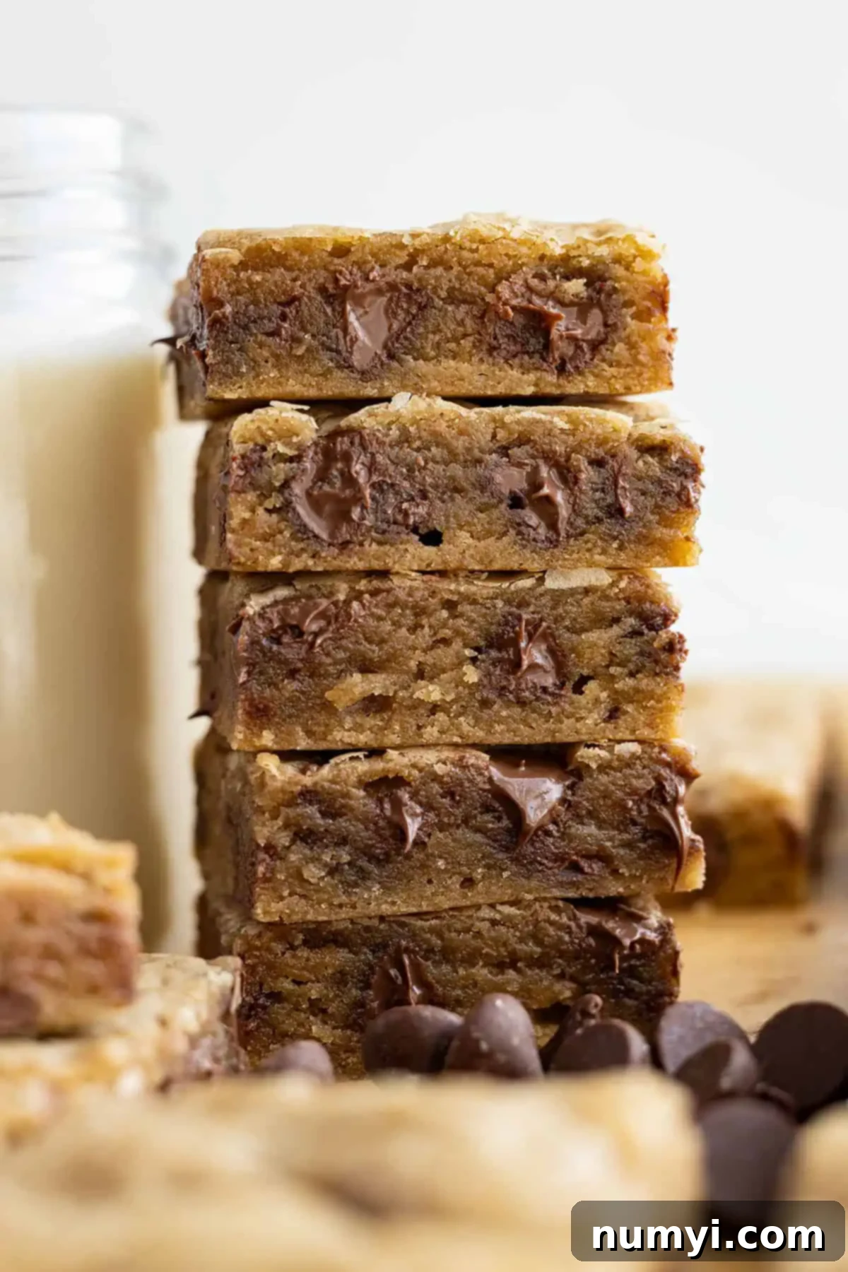 Tall stack of chocolate chip cookie bars next to a tall glass of milk, highlighting their soft and chewy texture.