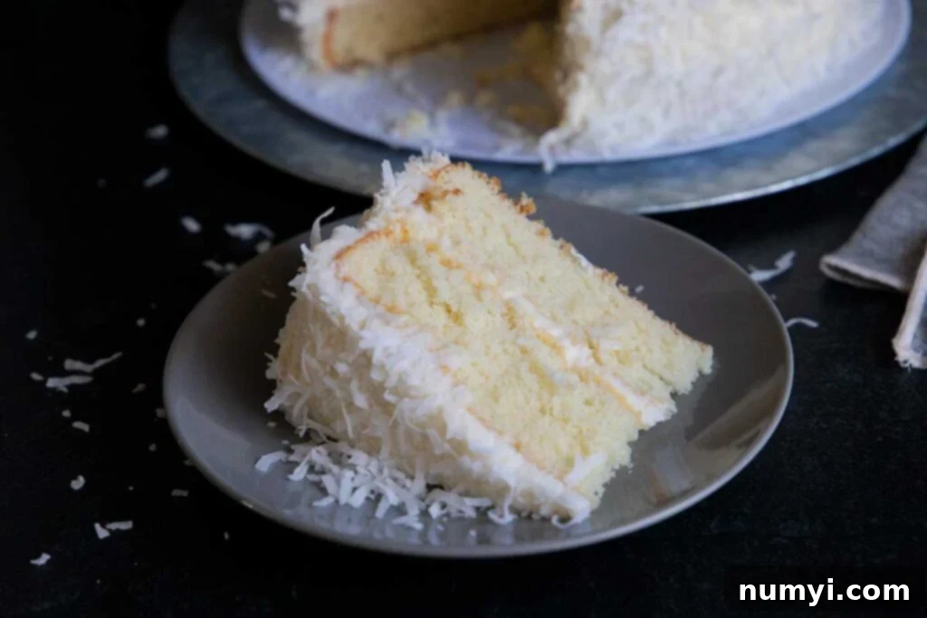 A delectable slice of homemade coconut cake, revealing its fluffy layers and creamy coconut buttercream frosting, rests on a light plate with the beautifully decorated whole cake visible in the soft-focus background.