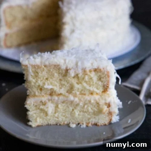 A beautifully sliced coconut cake, showcasing its moist, white layers generously frosted with coconut buttercream and adorned with sweet shredded coconut flakes, served on a grey plate.