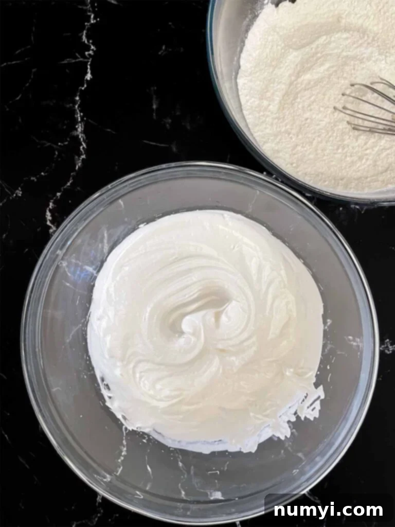 Perfectly whipped egg whites forming stiff peaks in a clear glass mixing bowl, ready to be incorporated into the coconut cake batter, set against a dark background.