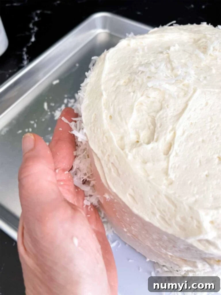 A hand gently pressing sweet shredded coconut flakes onto the sides of a freshly frosted coconut cake, creating a beautiful textured coating.