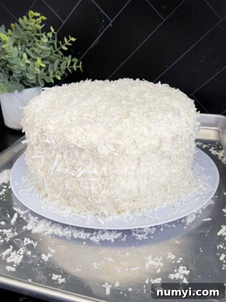 A perfectly frosted coconut cake, generously coated with shredded coconut, resting on a light plate atop a baking sheet, ready to be served.