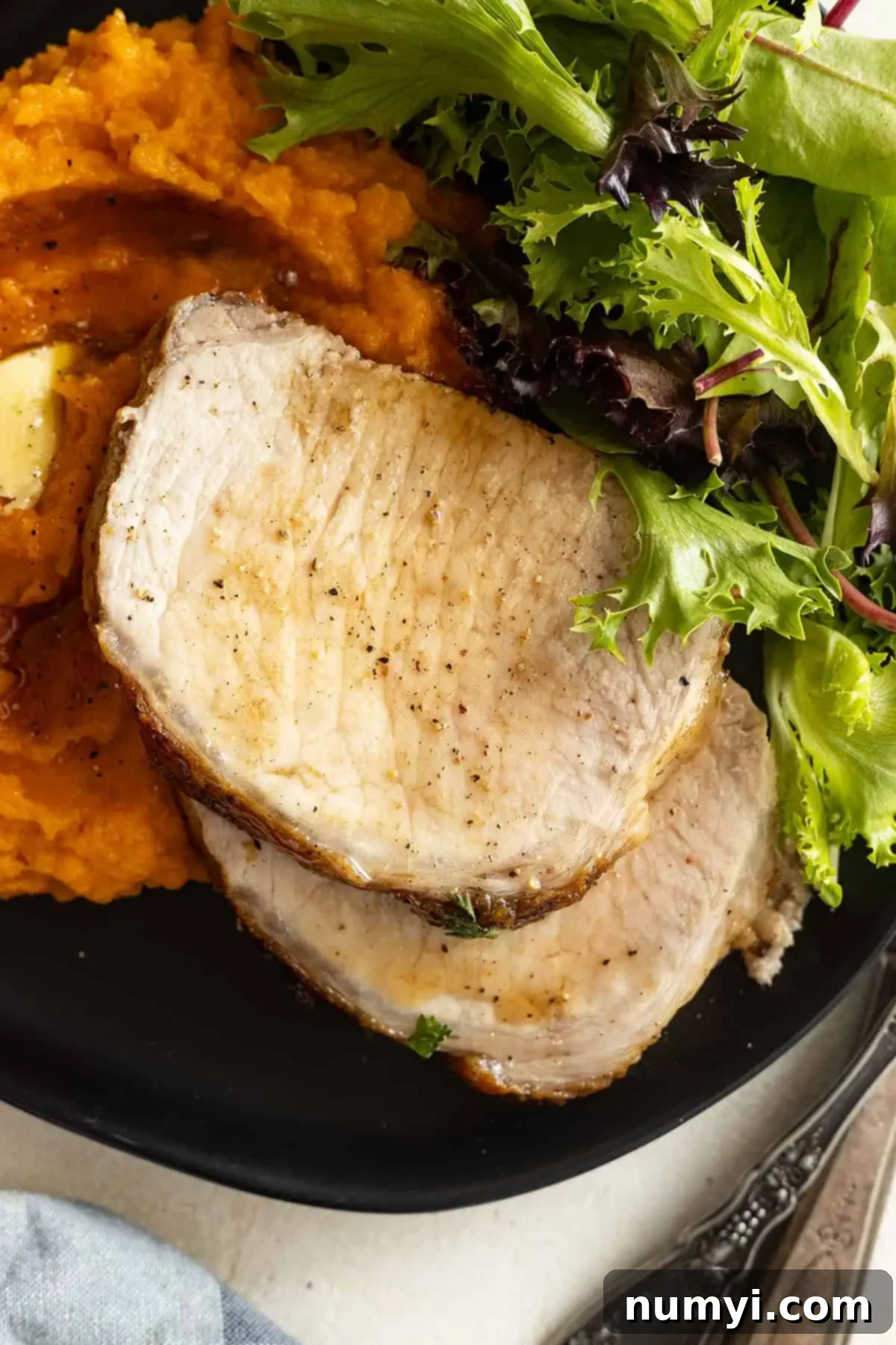Overhead view of perfectly sliced maple glazed pork roast on a black plate, served alongside fluffy mashed sweet potatoes and a vibrant green salad.