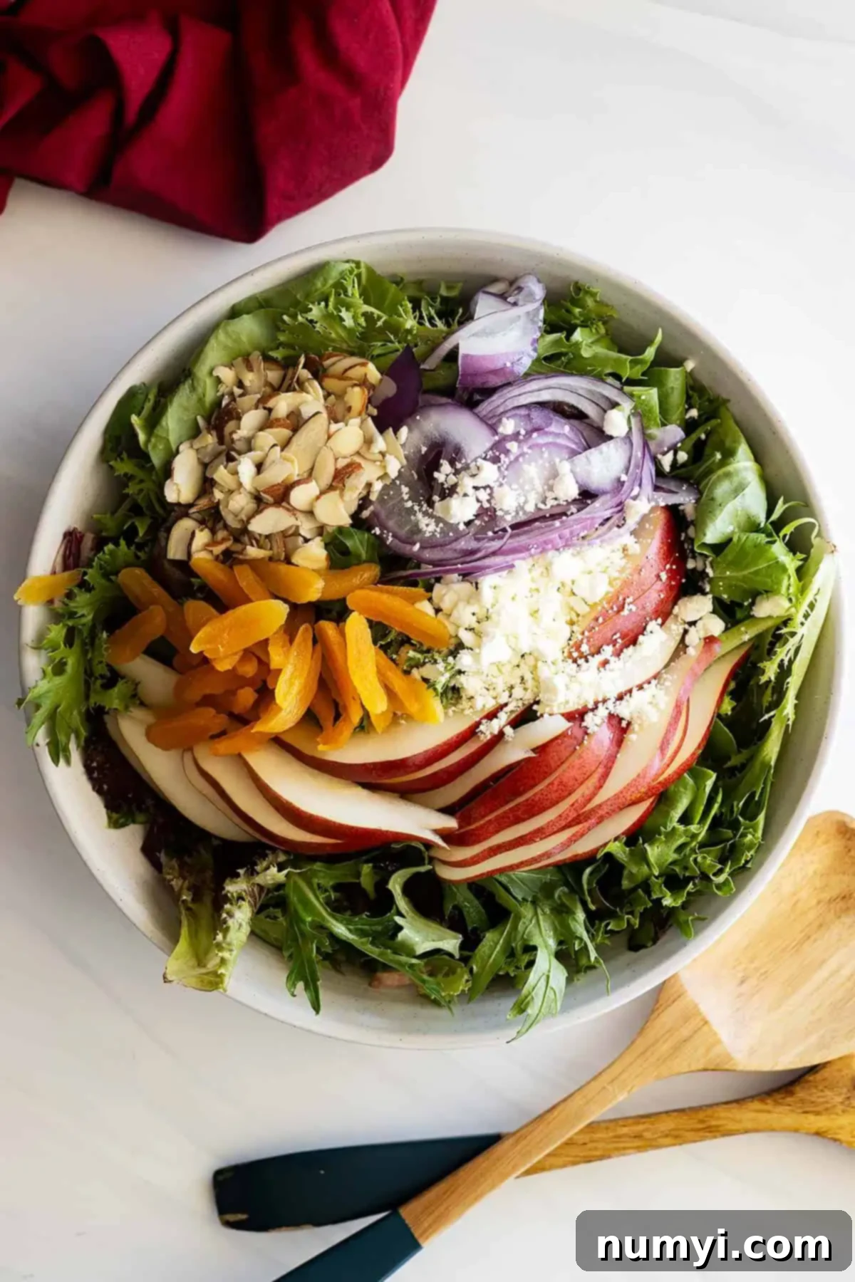 Overhead view of salad ingredients in a large white bowl, before tossing. Salad tongs and a red napkin are placed off to the side.