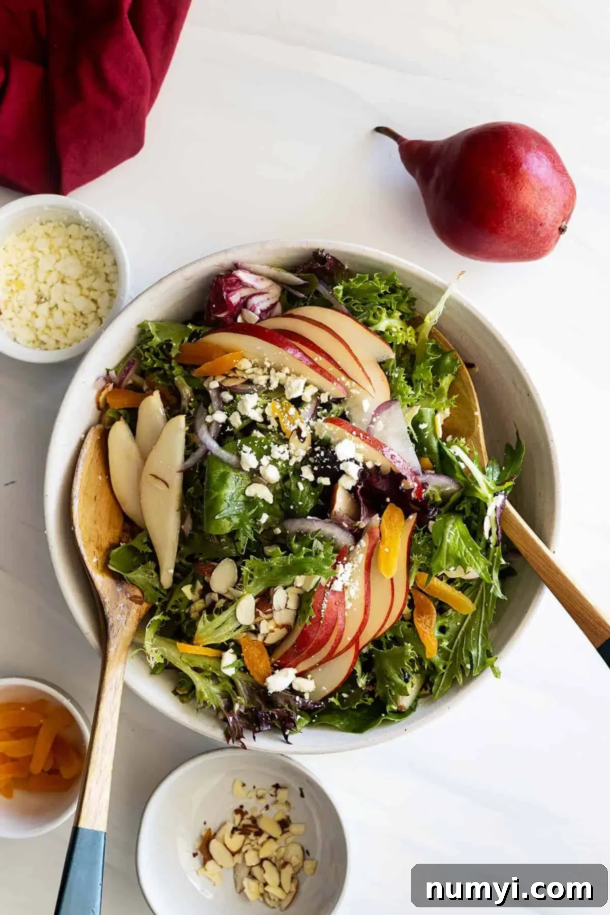 Overhead view of a perfectly tossed pear and apricot salad in a large white bowl, garnished with fanned pear slices and small bowls of its individual ingredients off to the side.