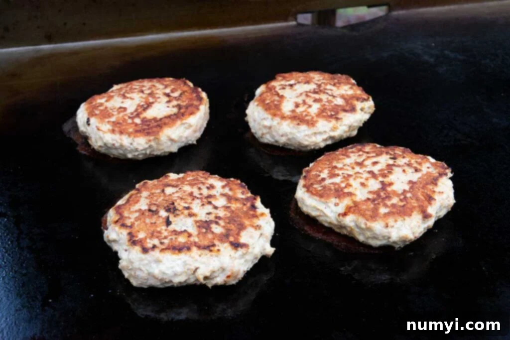 Close-up shot of four turkey burgers continuing to cook on a flat-top griddle, highlighting the browning process and savory sizzle.