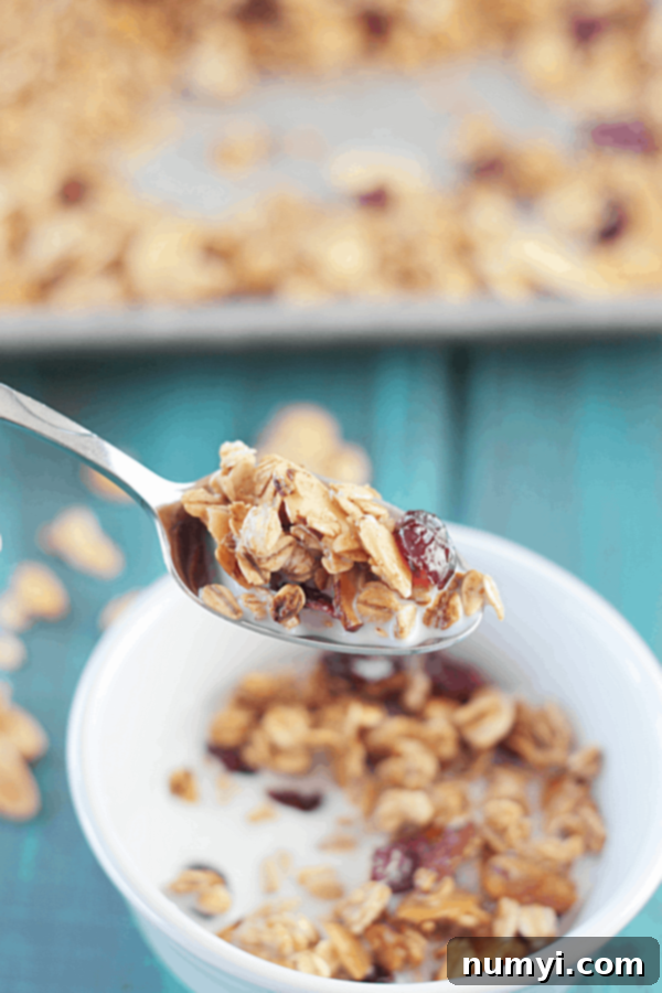 a scoop of homemade granola with maple and almond on a spoon with a bowl of granola and milk in the background