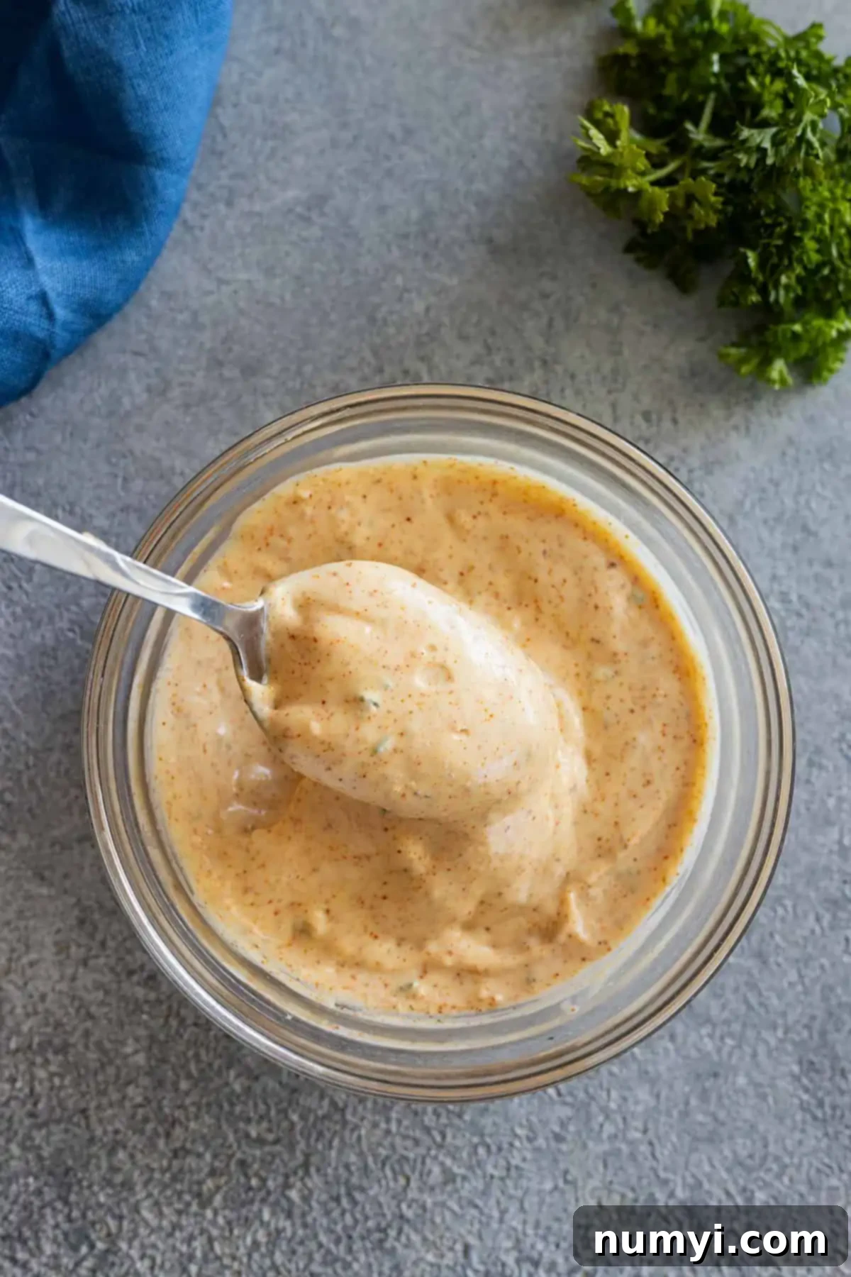 Overhead view of a bowl of remoulade sauce with a spoon dipping into it. Parsley and a blue napkin in the corners.
