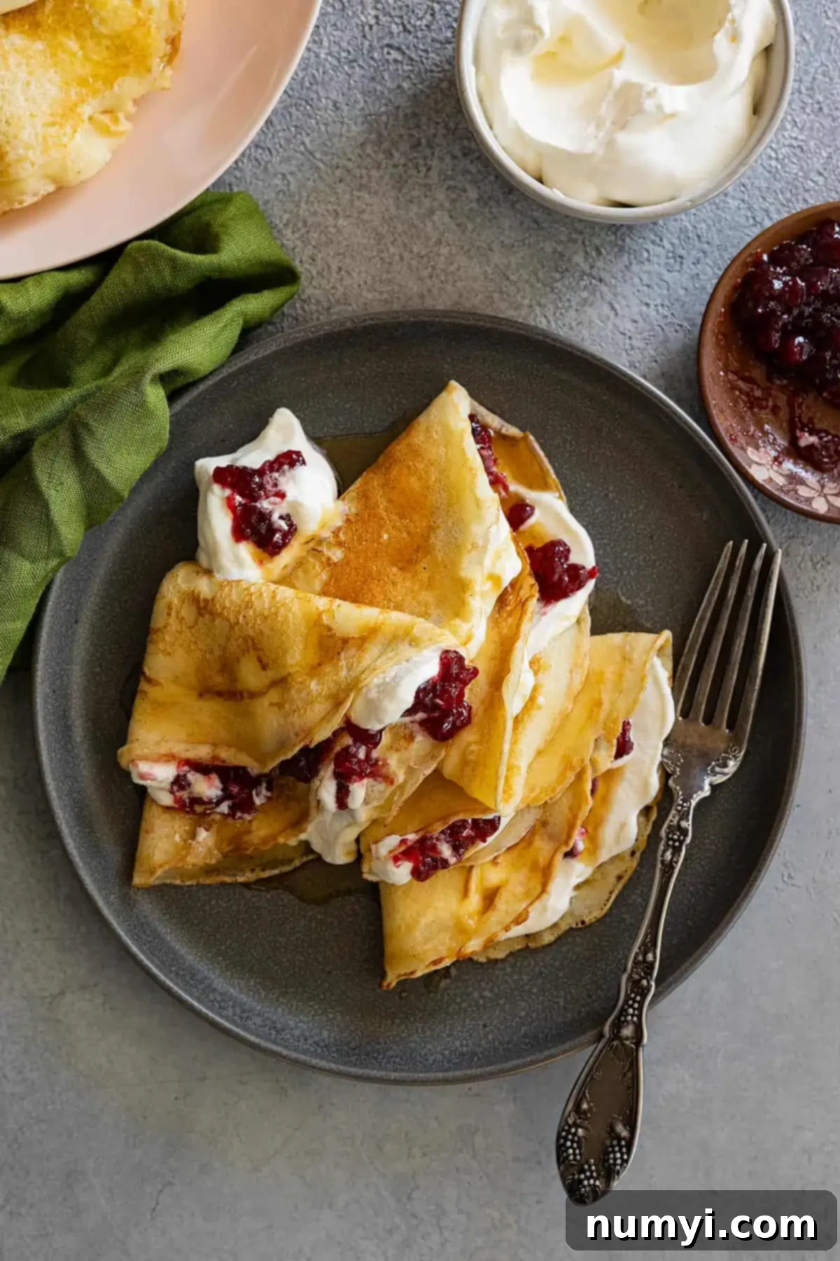 Overhead view of Swedish pancakes on a dark plate. They have whipped cream and lingonberries inside. Fork and green napkin to the side. 