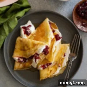 Overhead view of Swedish pancakes on a dark plate. They have whipped cream and lingonberries inside. Fork and green napkin to the side.