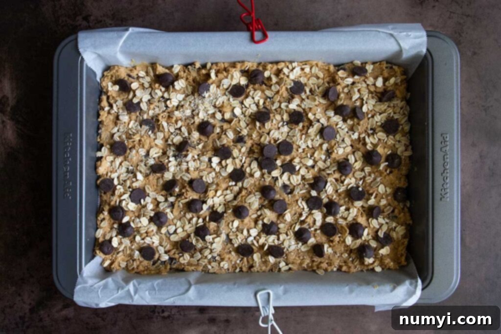 Oatmeal chocolate chip cookie bar dough spread evenly in a parchment-lined baking pan, ready for the oven.