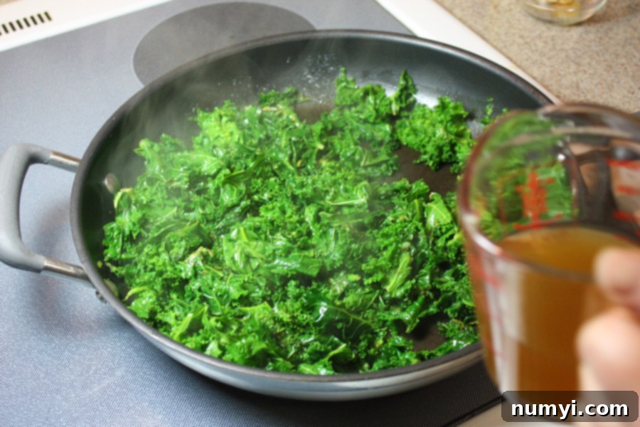 Chopped fresh kale leaves being sautéed in a large skillet with aromatic garlic and butter, showcasing the initial stages of the cooking process.