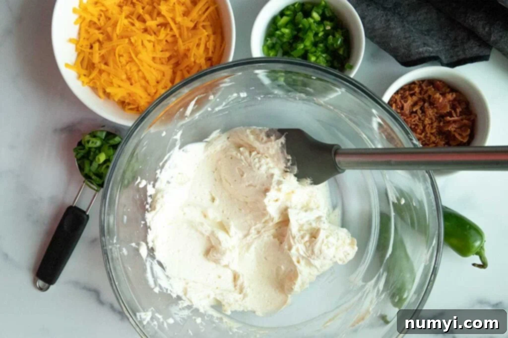 Softened cream cheese is prepared in a glass bowl, surrounded by neatly arranged shredded cheddar cheese, finely chopped jalapenos, vibrant diced green onions, and crispy chopped bacon, all ready to be combined for the cheese ball.