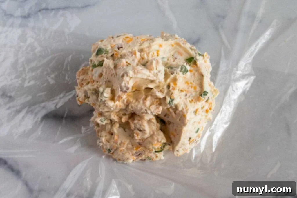 The Jalapeno Popper Cheese Ball, prior to its flavorful coating, is tightly wrapped in plastic wrap, ready for chilling.