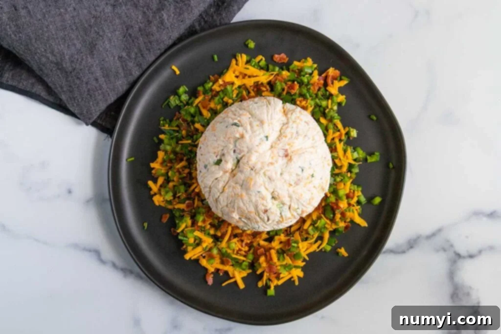 The chilled Jalapeno Popper Cheese Ball positioned atop its flavorful coating of bacon, jalapenos, and cheese on a dark plate, ready to be rolled.