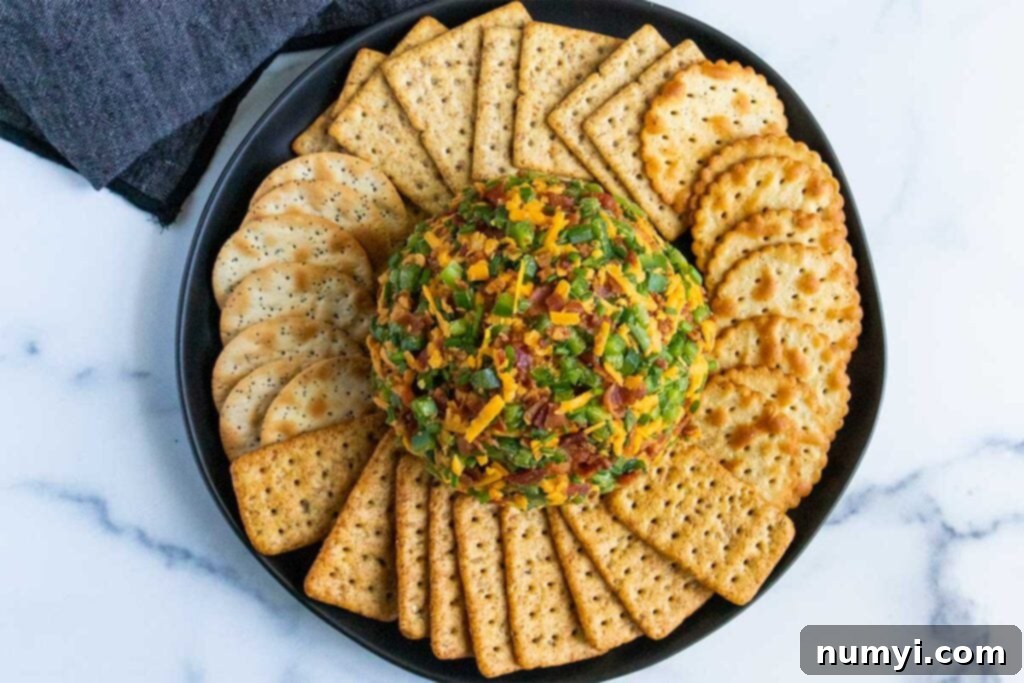 The fully coated Jalapeno Popper Cheese Ball, adorned with a generous layer of crispy bacon and diced jalapenos, is beautifully presented with an array of crackers on a dark plate, ready for serving.