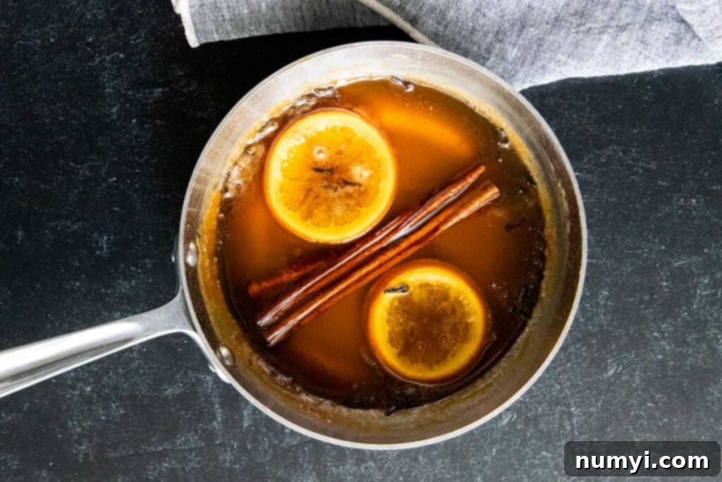 Honey syrup simmering and reducing in a medium saucepan, infused with orange slices, cinnamon sticks, and whole cloves.