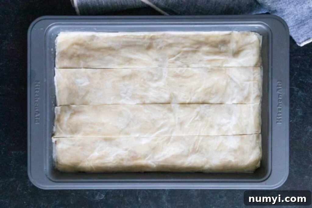 Initial straight cuts made into the unbaked baklava pastry in a baking pan, preparing for diamond shapes.