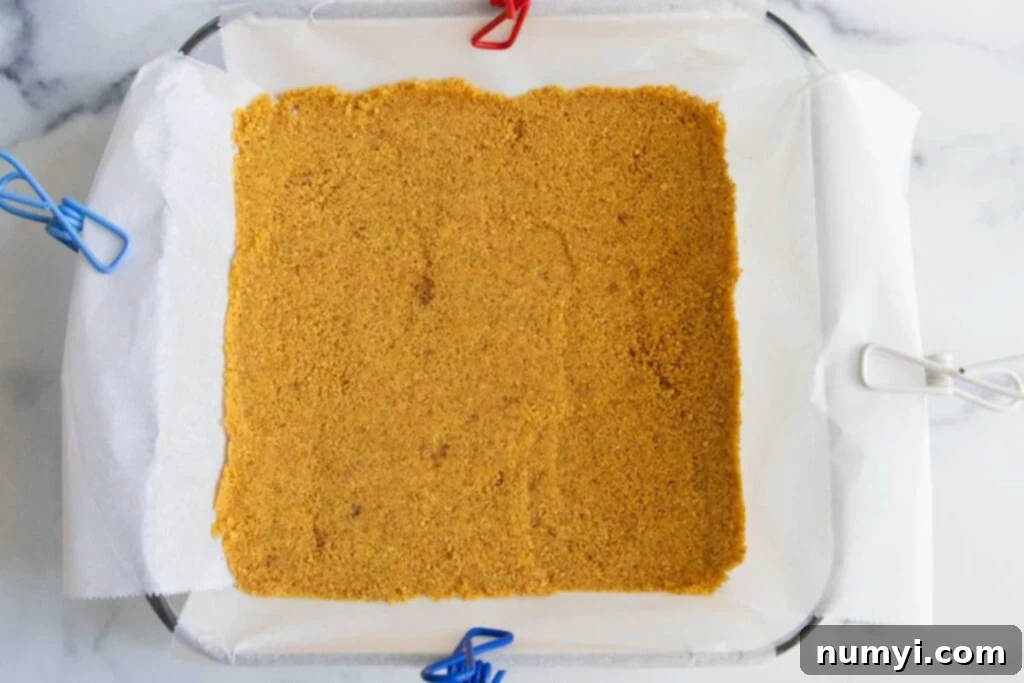 Golden brown graham cracker crust pressed evenly into the bottom of a parchment-lined baking dish, ready for the cheesecake layers.