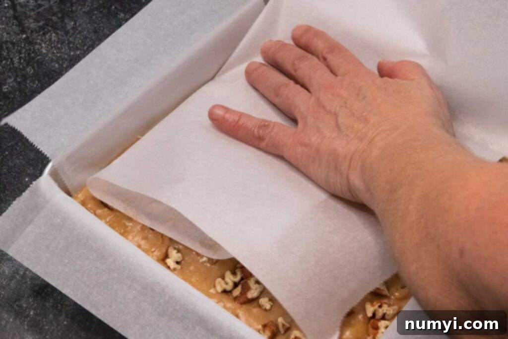 A hand gently pressing pecan pieces into the top of butter pecan fudge using parchment paper.