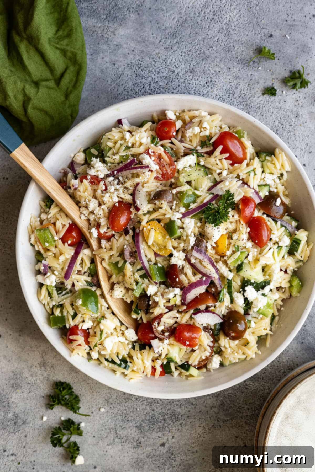 Overhead view of a vibrant Mediterranean orzo pasta salad in a white bowl with a spoon ready to serve, a green napkin artfully placed in the corner.