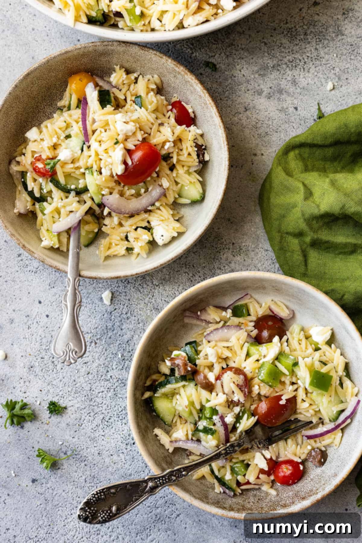 Two bowls with Mediterranean orzo pasta salad, each with a fork, ready to eat. A green napkin and bits of parsley are placed attractively on the sides.