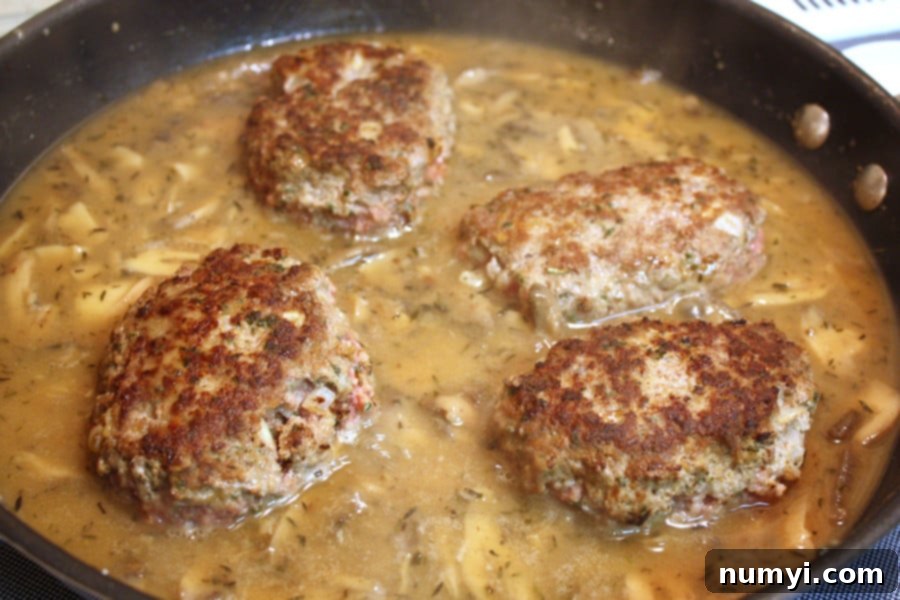 Homestyle Salisbury Steak with Rich Mushroom Gravy 3 Salisbury patties in the skillet with the gravy ingredients, showing the browned patties and mushrooms.