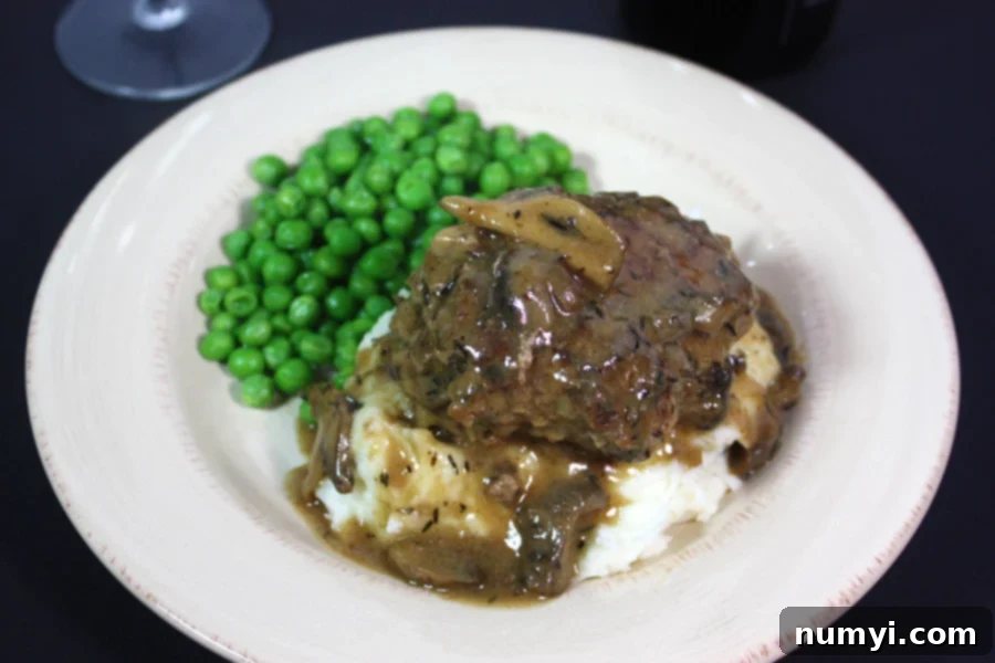 Homestyle Salisbury Steak with Rich Mushroom Gravy 5 A generous serving of Salisbury steak covered in gravy, nestled on a bed of mashed potatoes with a side of green peas on a white dinner plate.