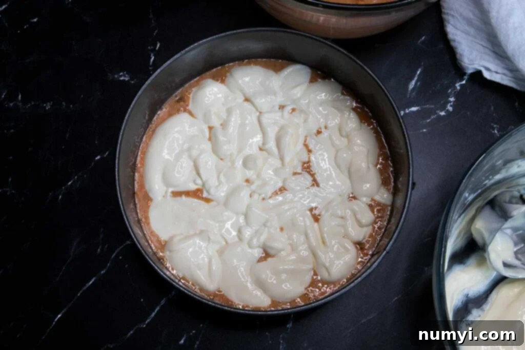 A layer of creamy cheesecake mixture carefully dolloped over the carrot cake batter in a springform pan, creating distinct layers.