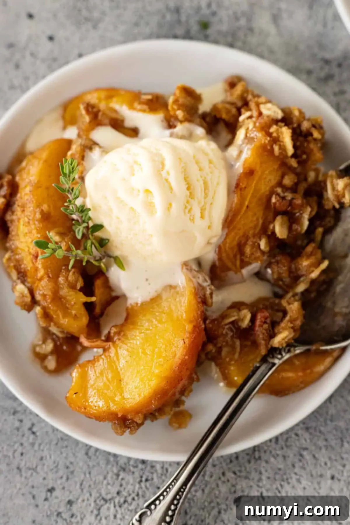 Overhead view of a golden-brown peach crisp on a white plate with vanilla ice cream slowly melting into the warm dessert.