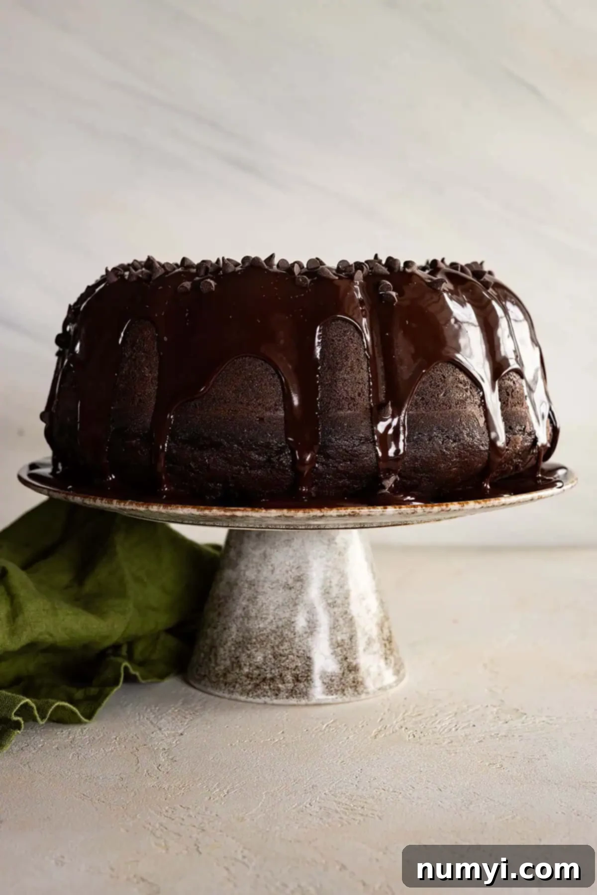 A perfectly baked and glazed Chocolate Zucchini Bundt Cake presented on an elegant cake stand, with a decorative green napkin to the side.