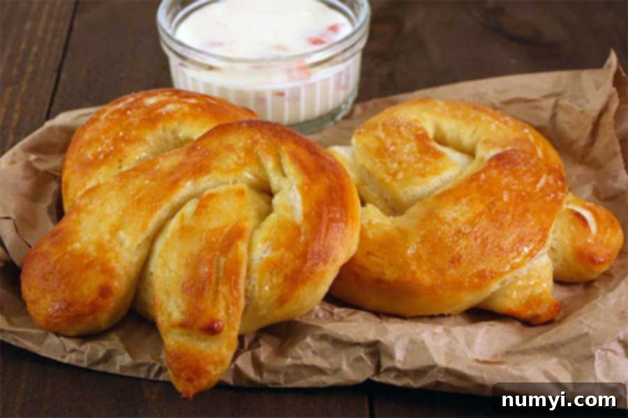 A stack of golden-brown soft pretzels cooling on brown paper, with a ramekin of cheese dip blurred in the background, ready for dipping.
